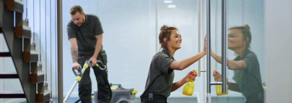 Two janitors cleaning: one is vacuuming the floor; the other is cleaning a glass wall with a spray bottle and cloth. Both are wearing dark uniforms.
