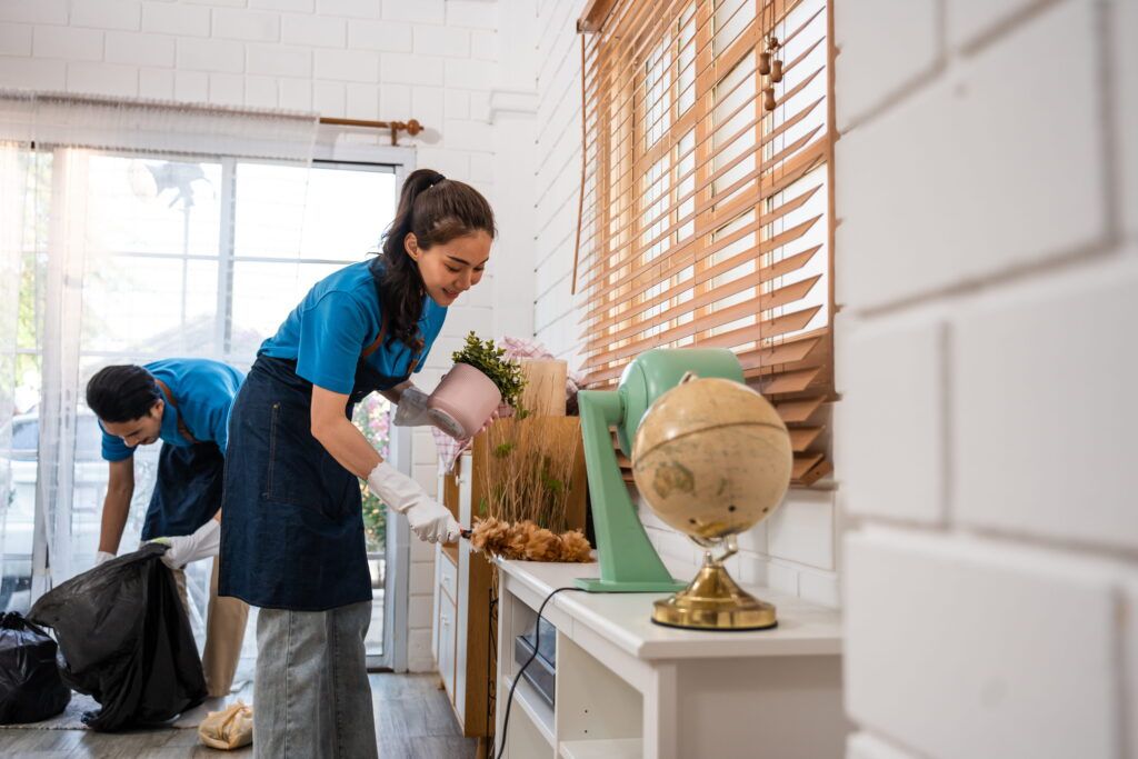Two people wearing blue shirts and aprons are cleaning a room; one is dusting a shelf while the other collects trash in a black bag.