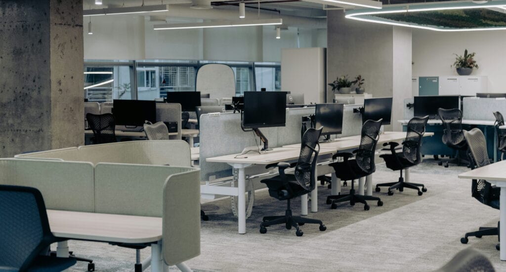 A modern open-plan office with rows of desks, multiple computer monitors, office chairs, and minimal decor, with large windows in the background.