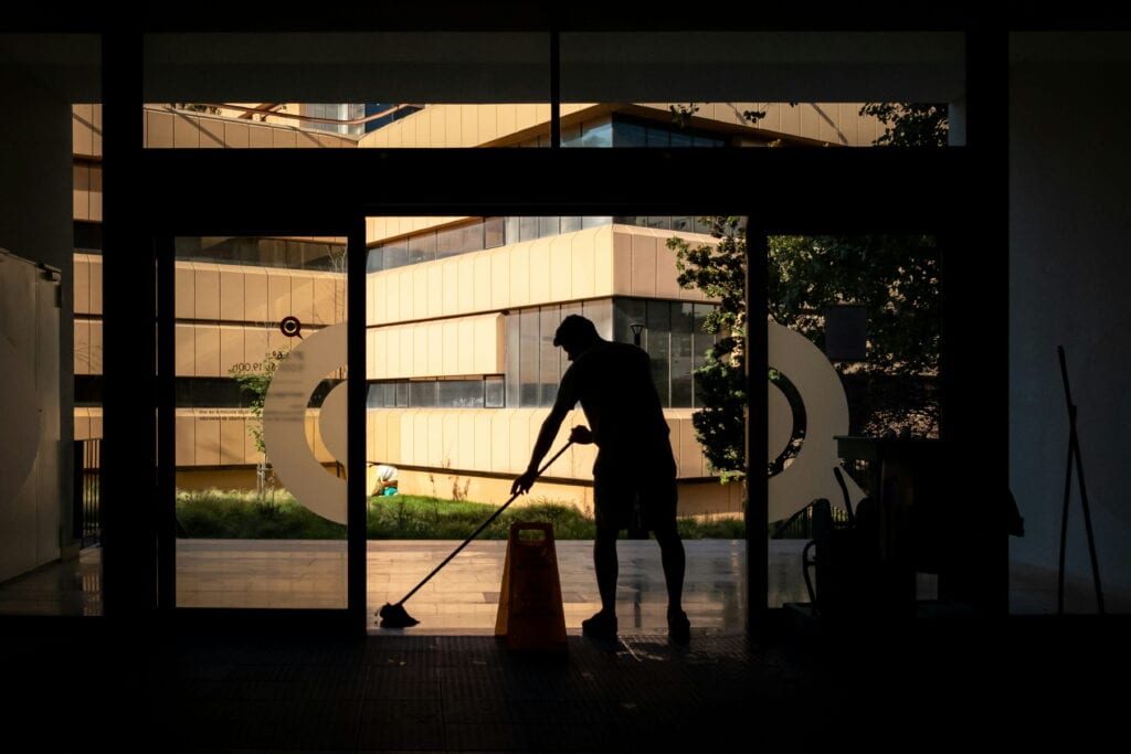 A person mops the floor inside a building near a large window, with a caution wet floor sign placed nearby and modern buildings visible outside.