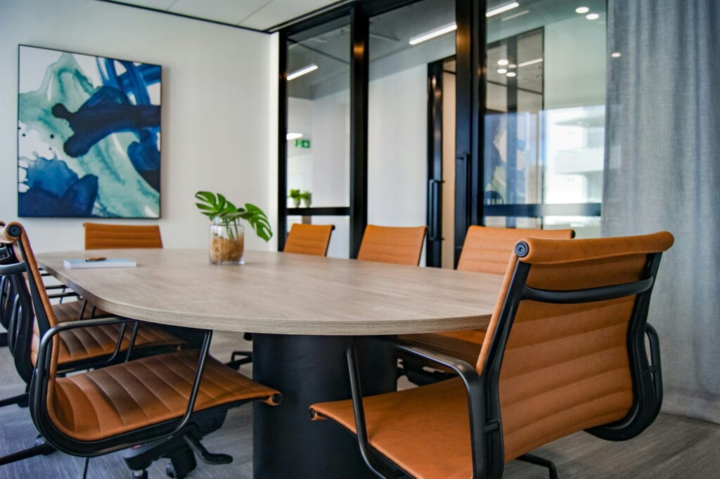 A modern conference room with a wooden table, orange chairs, a plant centerpiece, glass walls, and abstract art on the wall.