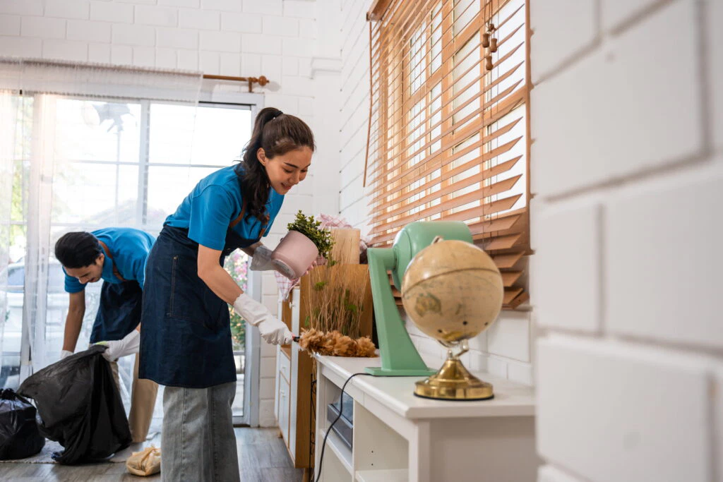 Two people wearing blue shirts and aprons are cleaning a room; one is dusting a shelf while the other collects trash in a black bag.
