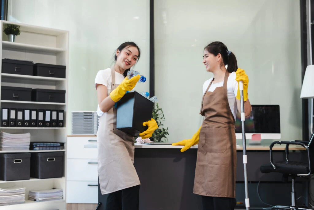 Two women in aprons and yellow gloves clean an office; one discards bottles into a bin, the other holds a mop.