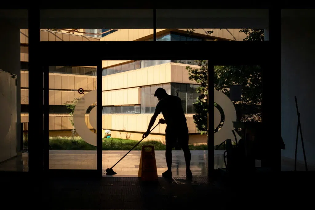 A person mops the floor inside a building near a large window, with a caution wet floor sign placed nearby and modern buildings visible outside.