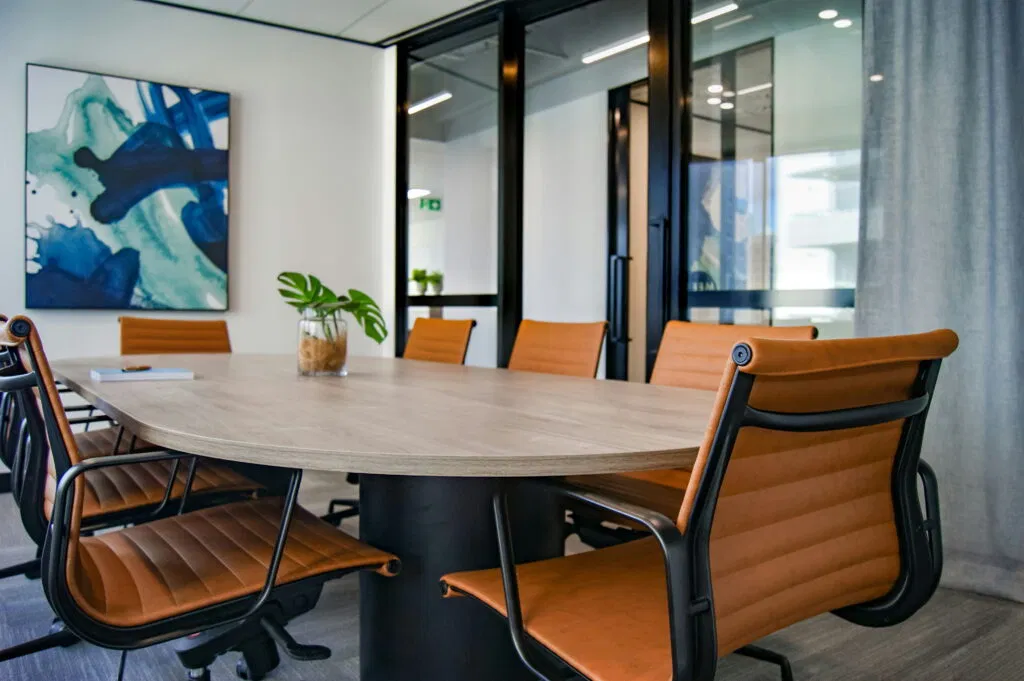 A modern conference room with a wooden table, orange chairs, a plant centerpiece, glass walls, and abstract art on the wall.